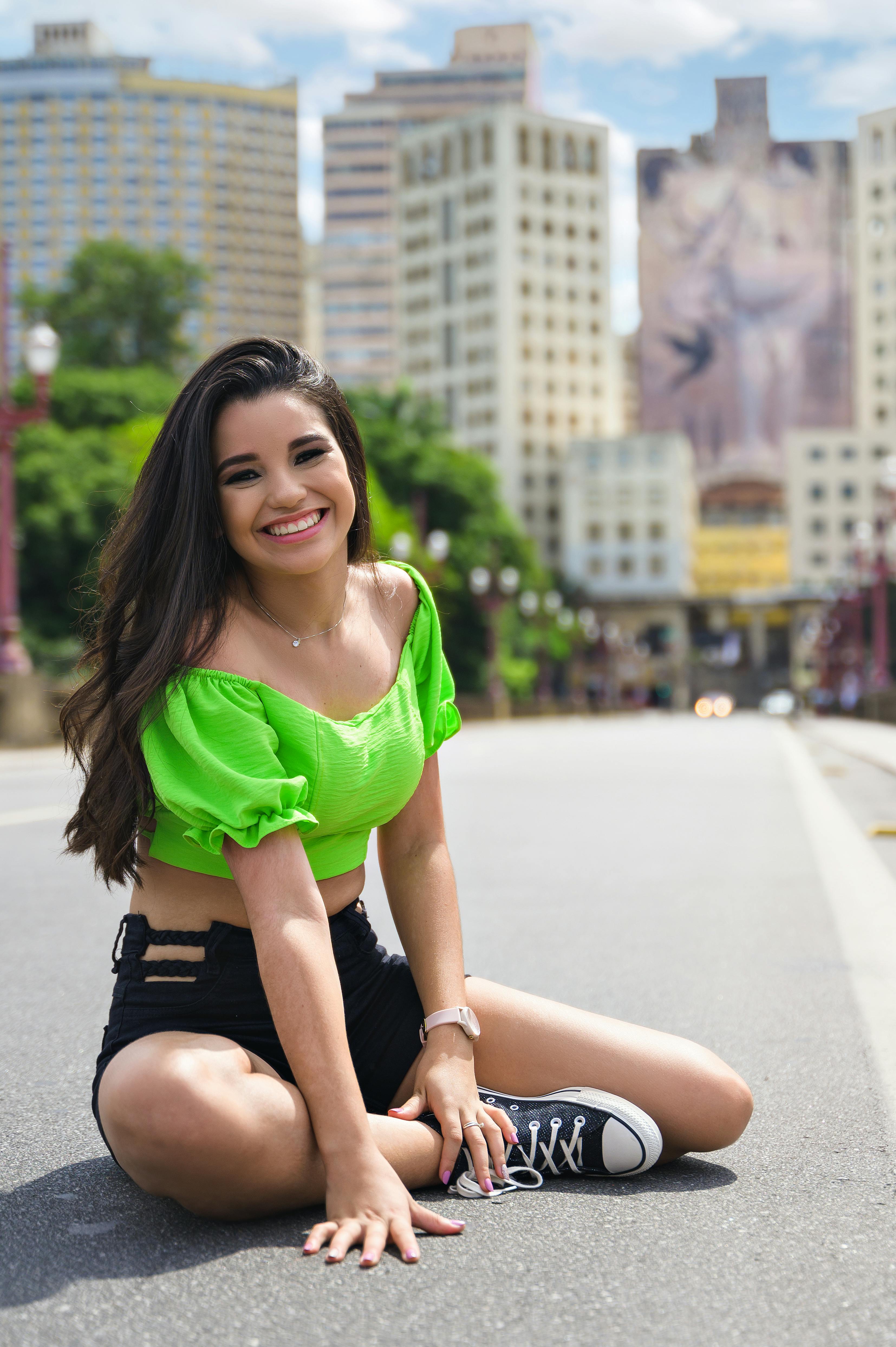 Happy young woman in green top sits on urban street, smiling with buildings in the background.