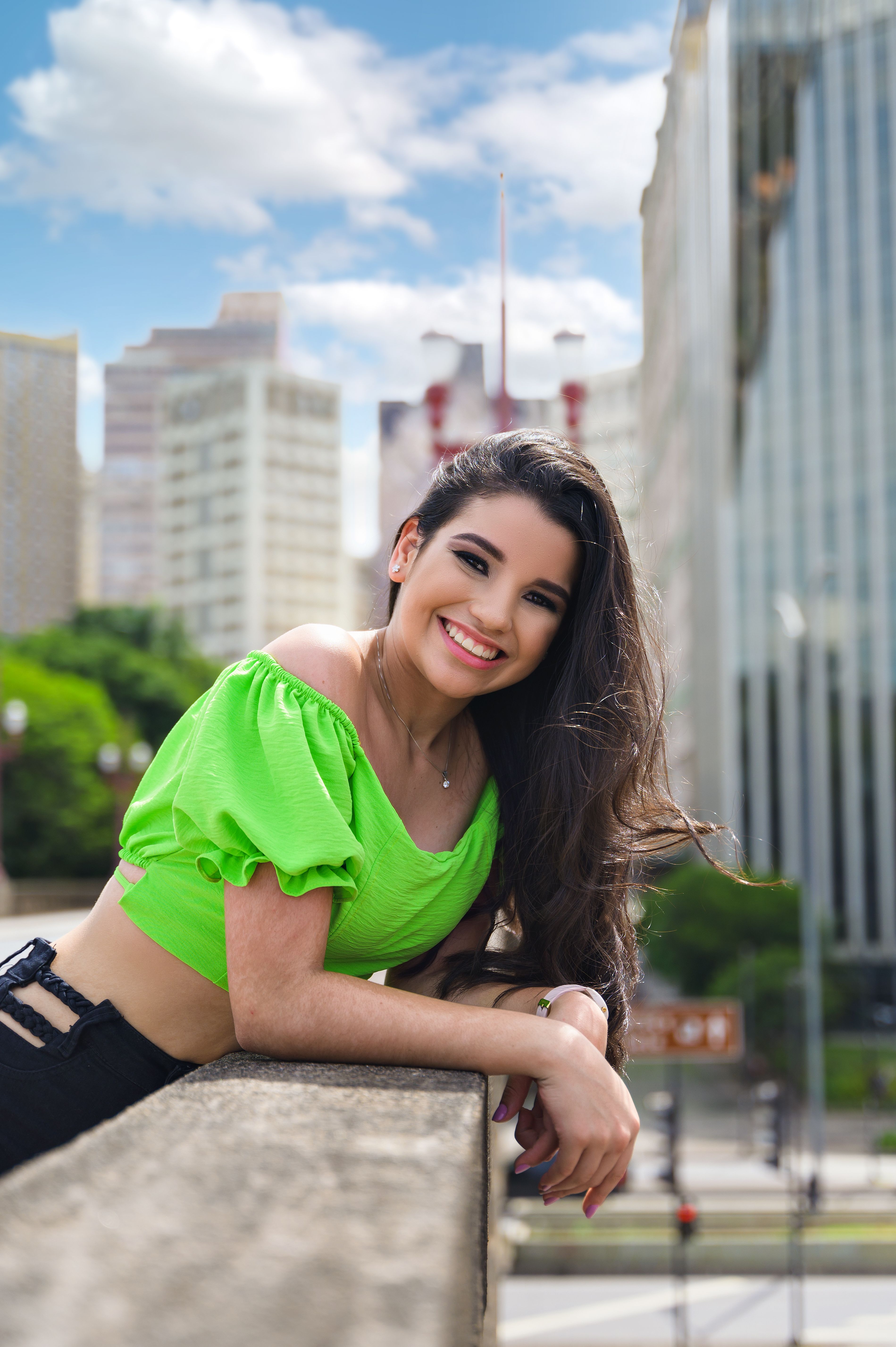 A young woman in a green crop top smiles on a rooftop with a cityscape background.