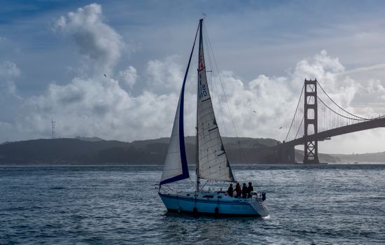 A sailboat navigating near the iconic Golden Gate Bridge in San Francisco under a cloudy sky.