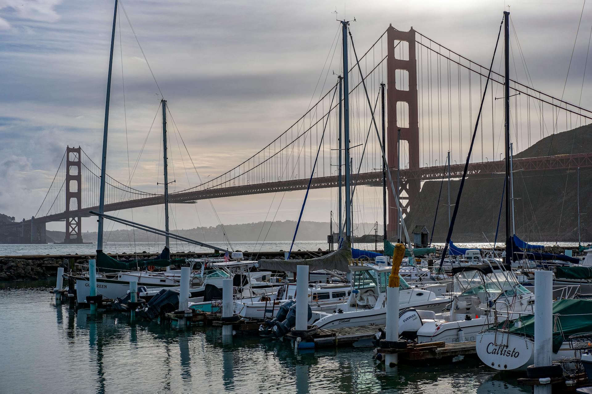Sailboats docked at a marina with the iconic Golden Gate Bridge silhouetted against a sunset sky.