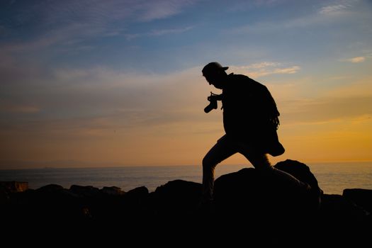 Silhouette of a person with a camera on coastal rocks during sunset, capturing scenic sea views.