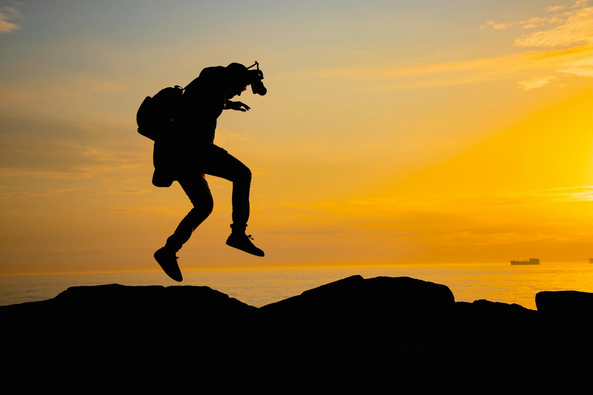 A man silhouetted against a vibrant sunset sky, jumping over rocks by the sea.