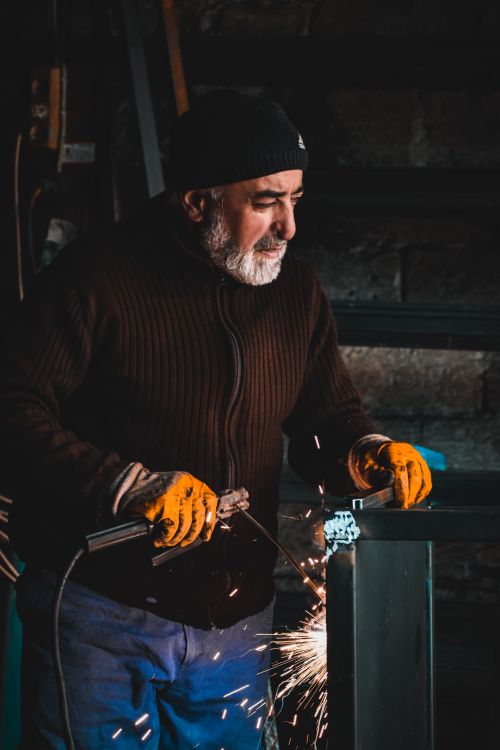 Free Elderly man welding with gloves and hat in a workshop, generating bright sparks. Stock Photo