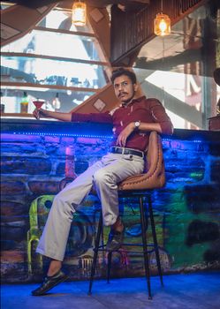A fashionable man in a maroon shirt relaxing at an urban bar with a drink in hand.