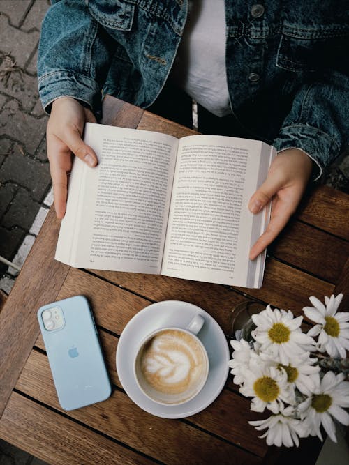 Person Holding a Book Beside a Cup of Cappuccino