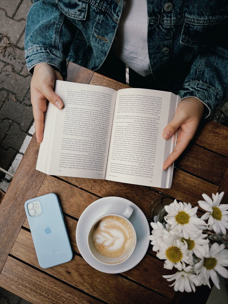 Person Holding A Book Beside A Cup Of Cappuccino