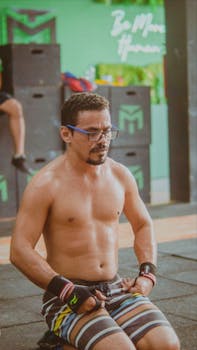 A fit, shirtless man with glasses kneels in a gym, focusing on his workout with determination.