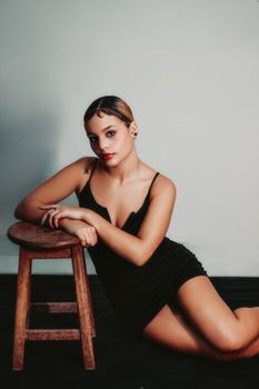 Elegant portrait of a woman in black dress posing next to a wooden stool indoors.