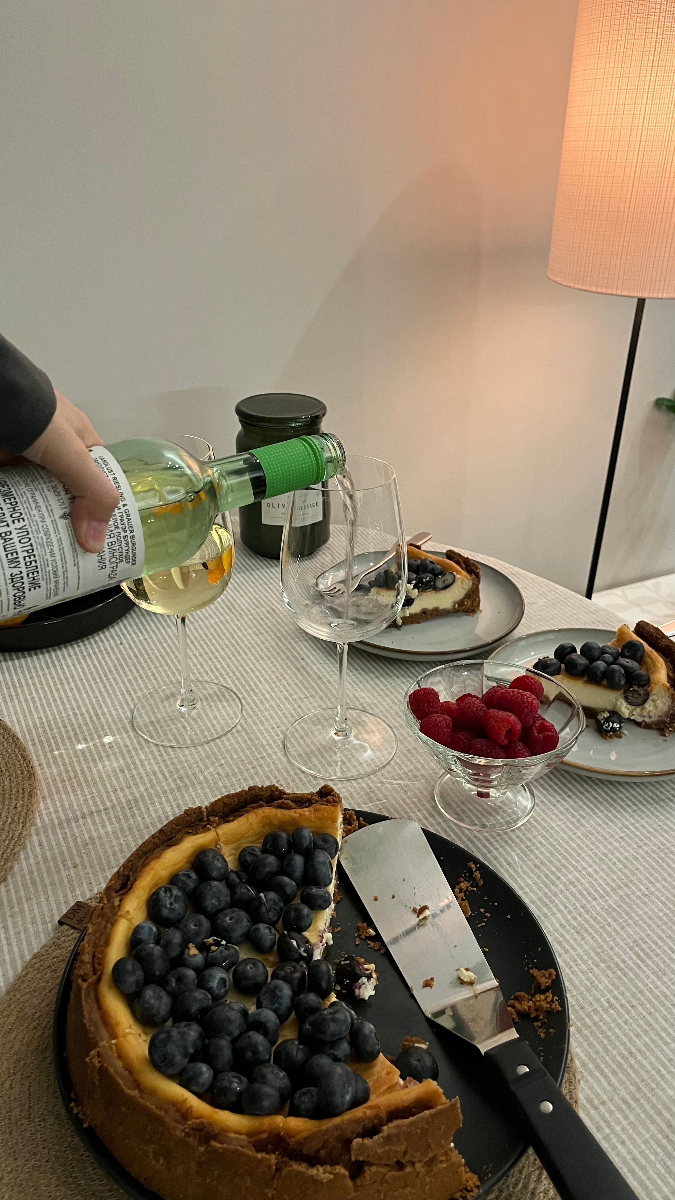 A wine bottle pouring into glasses beside blueberry cheesecake and raspberries on a table.
