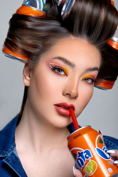 Artistic portrait of a woman with soda cans in hair, sipping from a can with a straw.