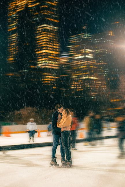 A romantic couple shares a kiss while ice skating outdoors on a snowy winter night in the city.