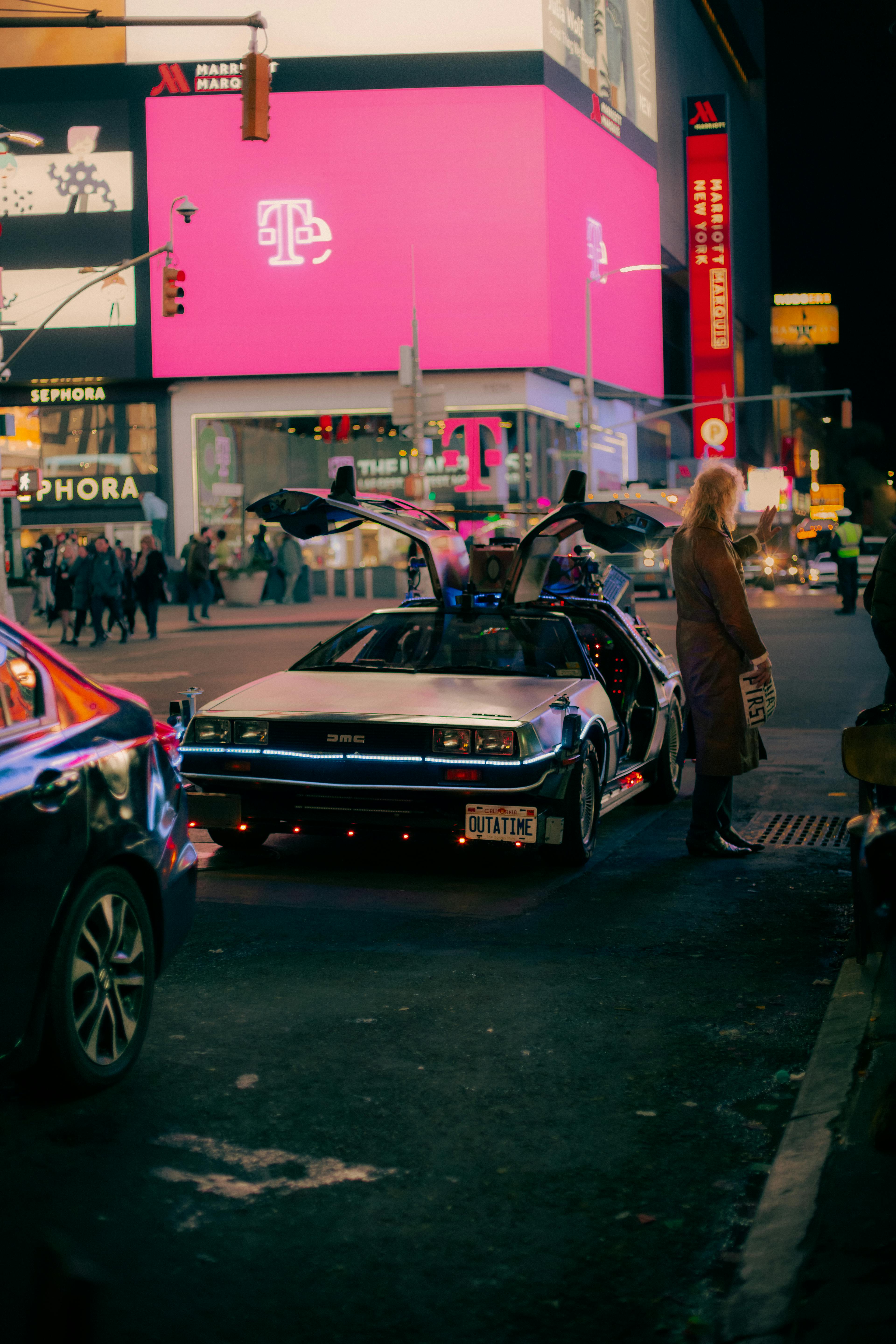 A classic DeLorean DMC car parked on a bustling city street at night. The scene is vibrant with urban energy.