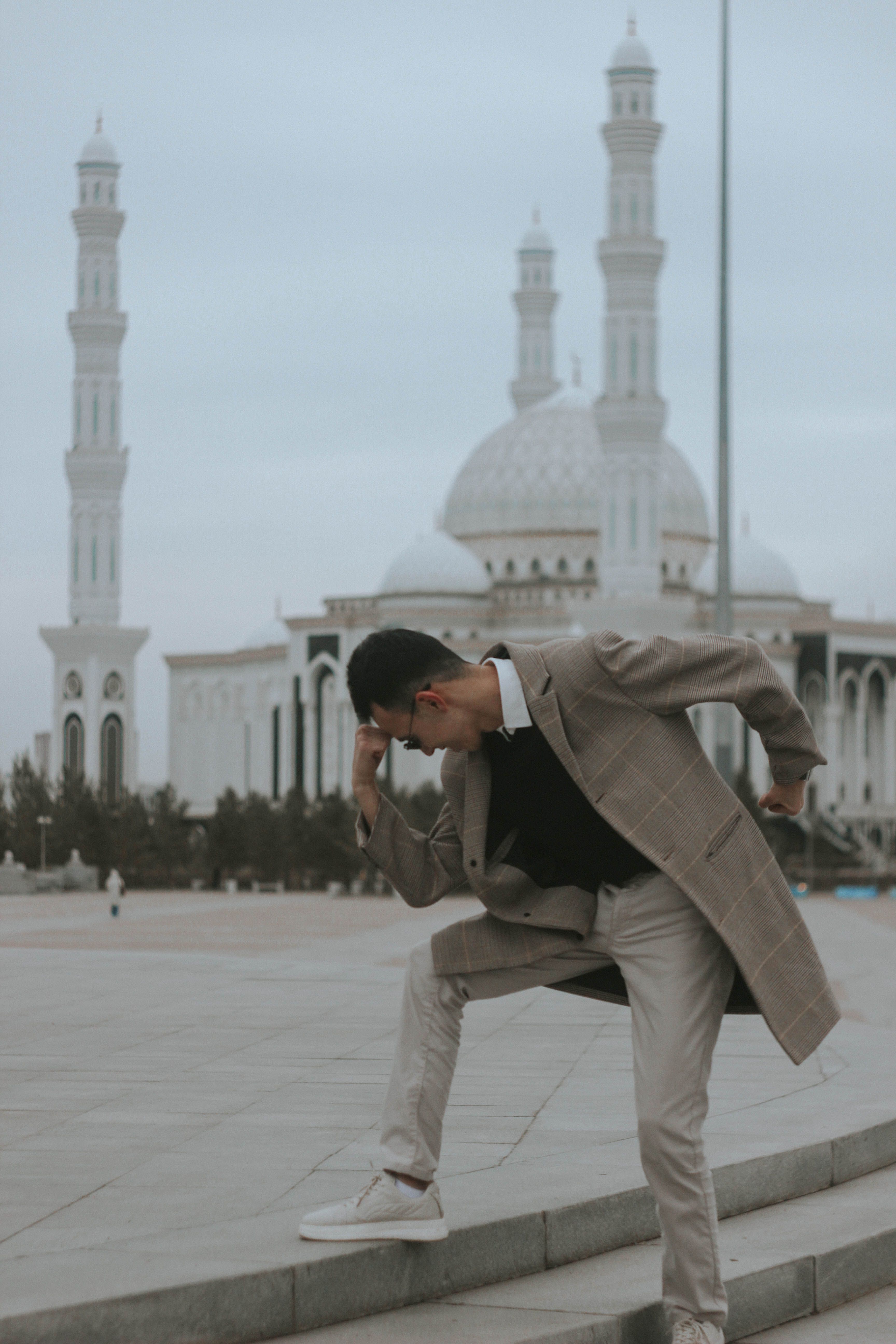 Stylish man in a coat posing creatively in front of a grand mosque with towering minarets.