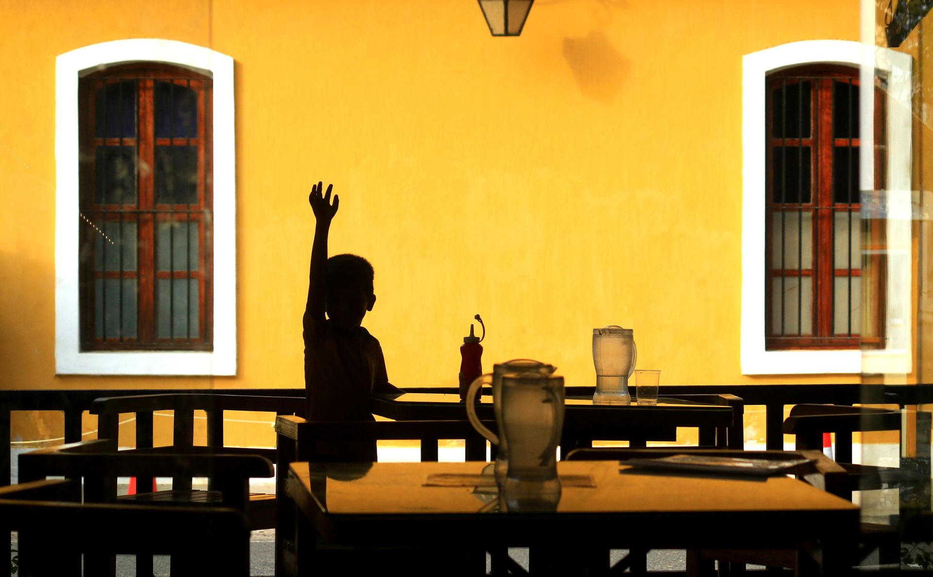 Silhouette of a child raising hand at outdoor patio with vibrant yellow wall and windows.