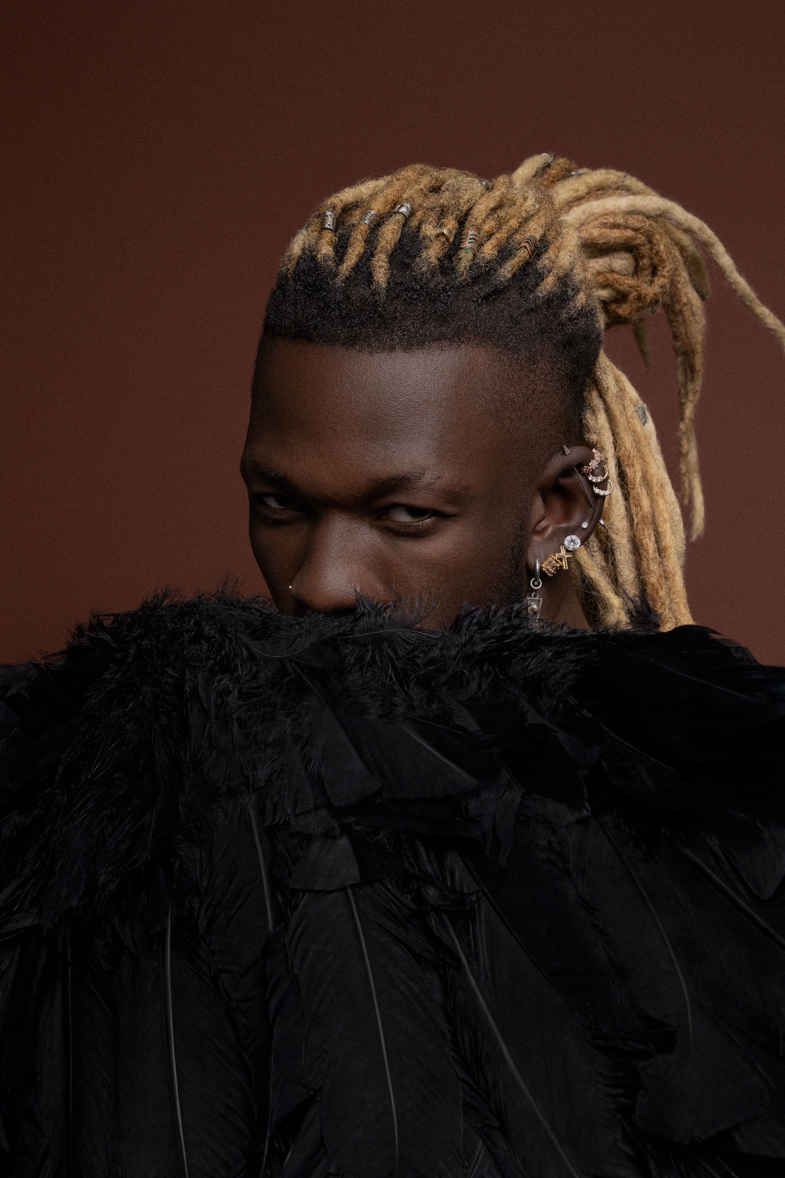 Fashion-forward portrait of a man with blonde dreadlocks and a feathered outfit against a brown background.