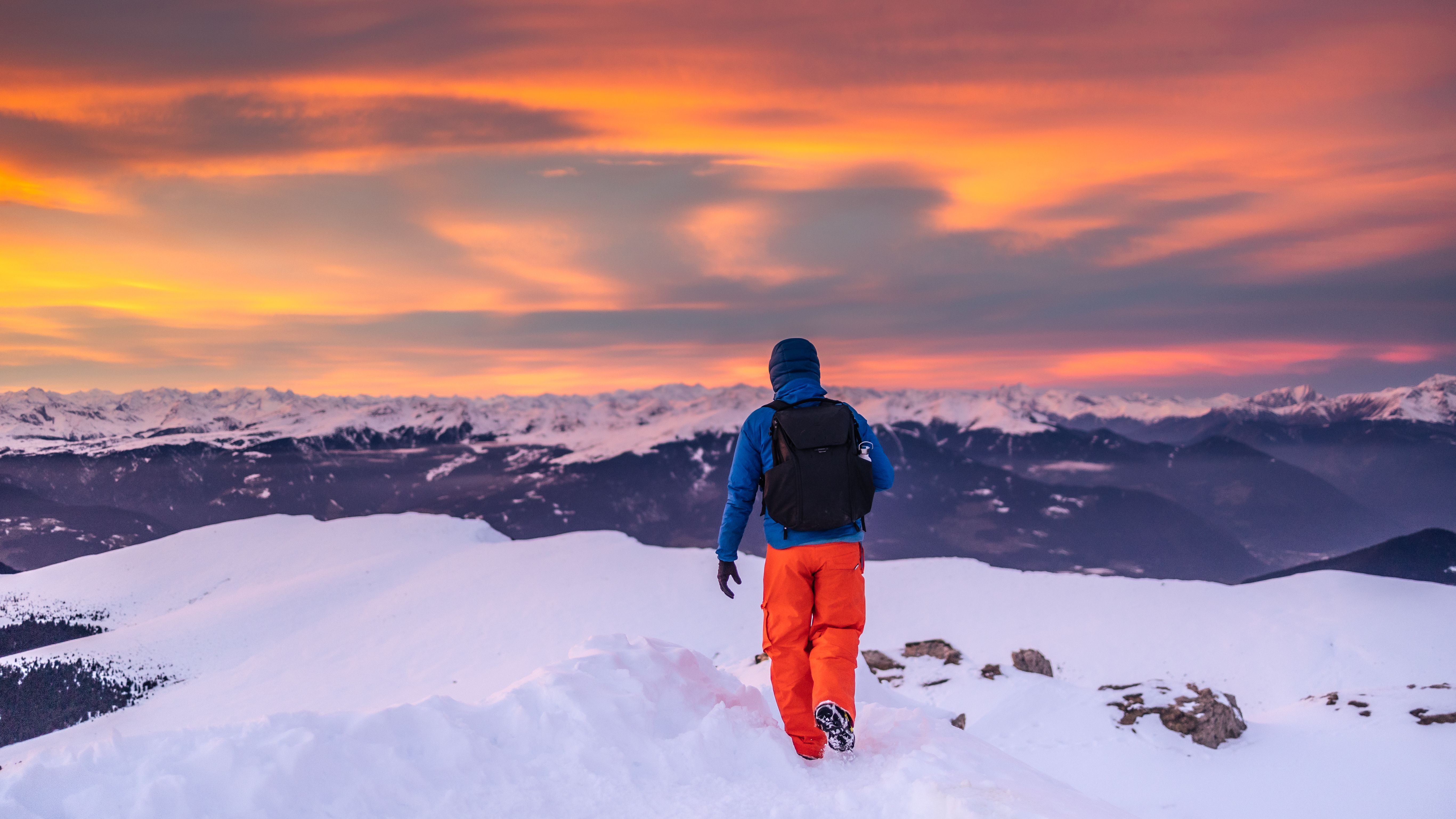 Man in winter gear hiking in snowy mountains under a vibrant sunset sky