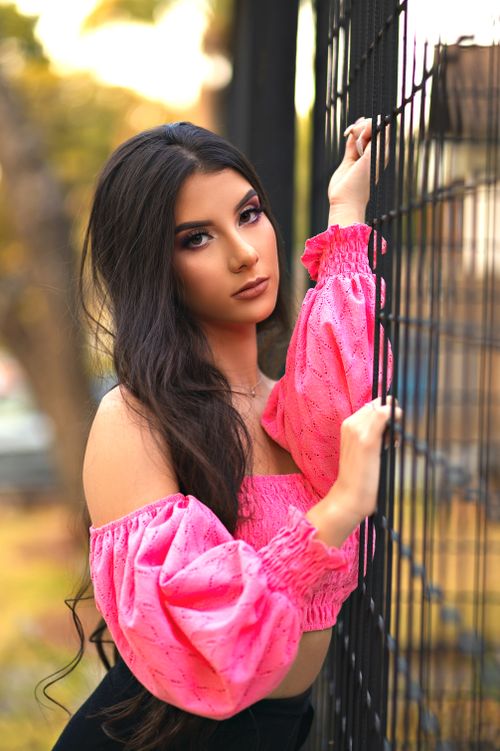 Free A young woman in a pink off-shoulder top poses by a fence outdoors in a vibrant, sunny setting. Stock Photo