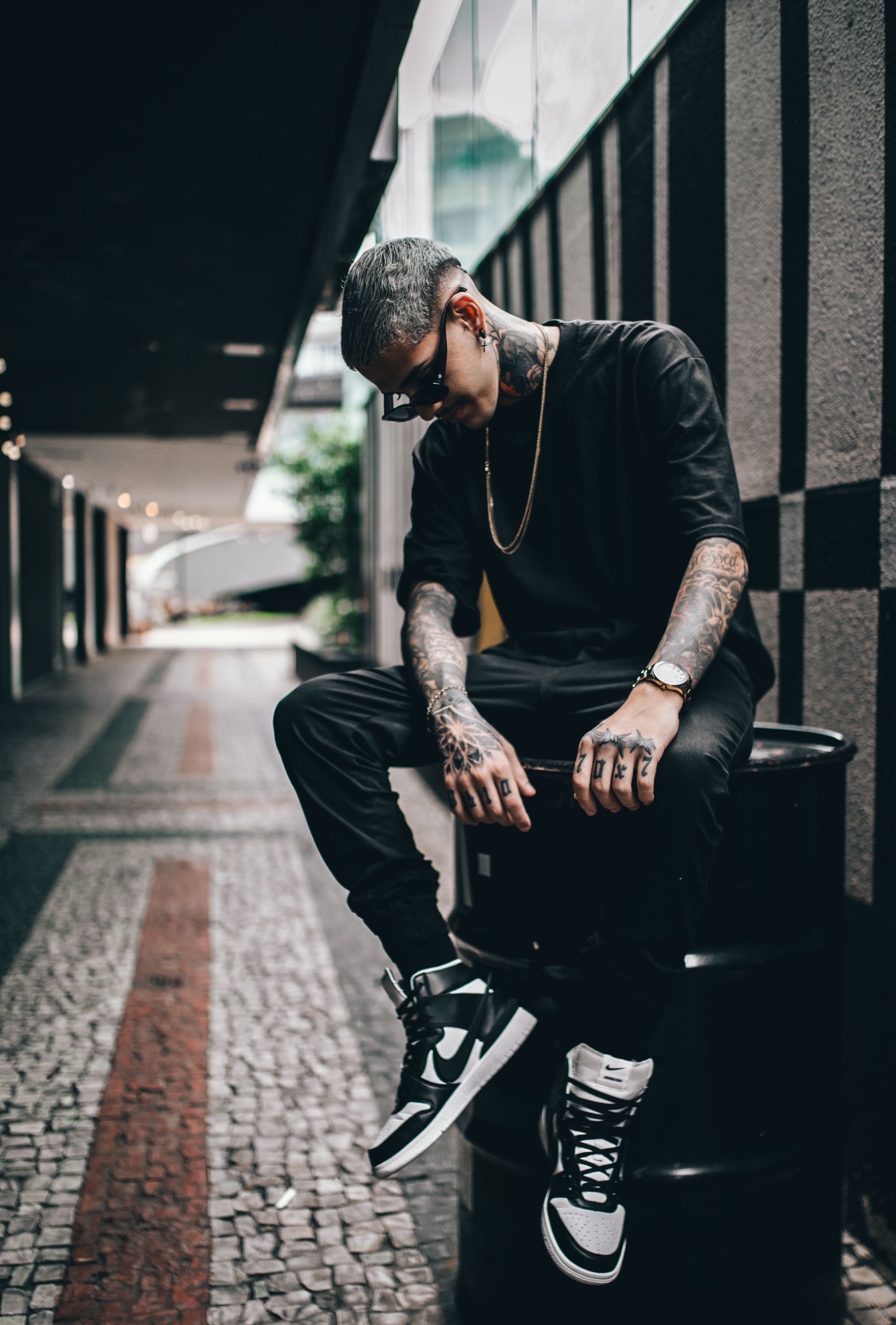 Um homem tatuado em estilo streetwear sentado em um barril em um beco urbano, exalando um estilo moderno e descolado.