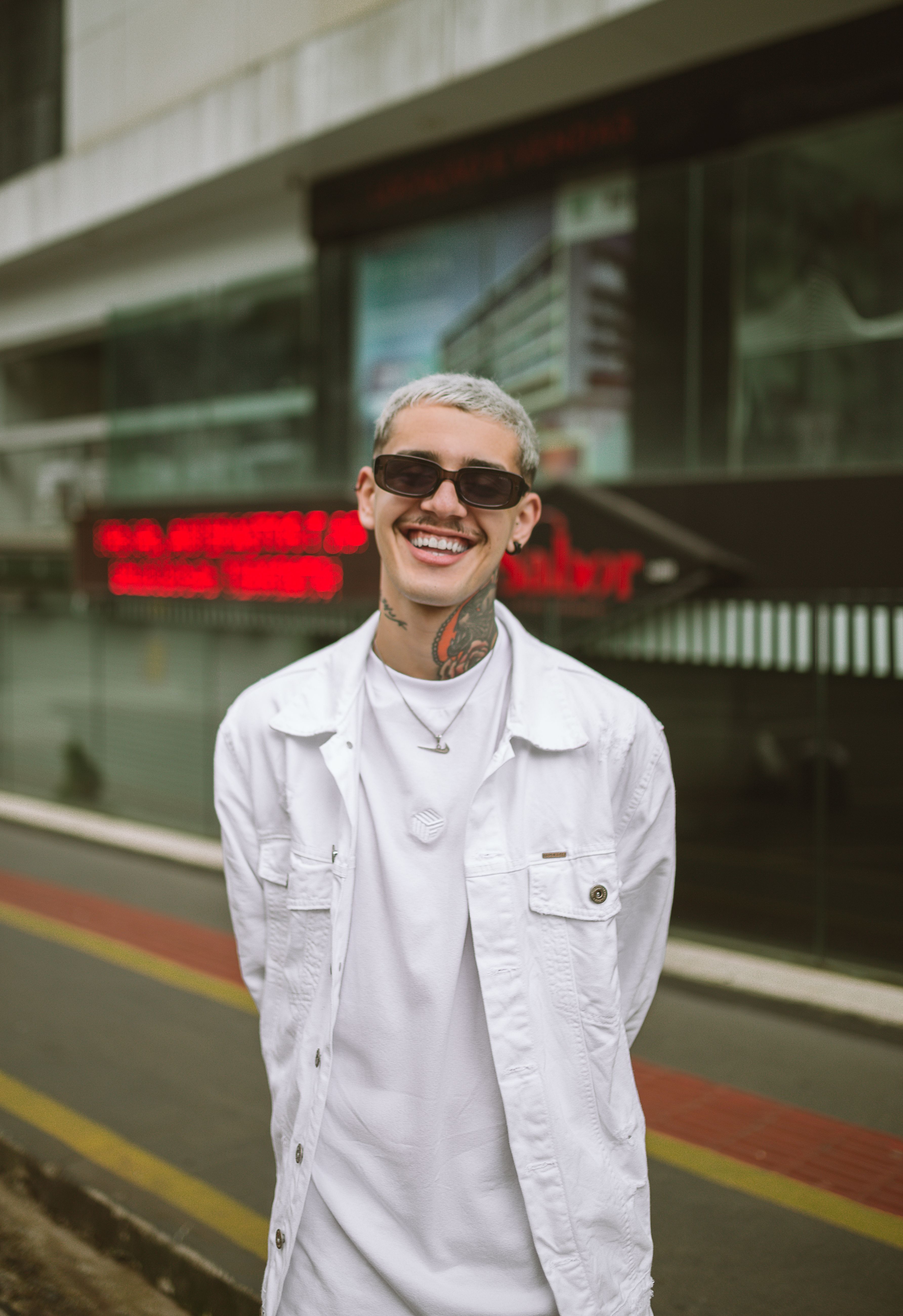 A stylish man with sunglasses and tattoos smiling on a city street, exuding confidence and happiness.