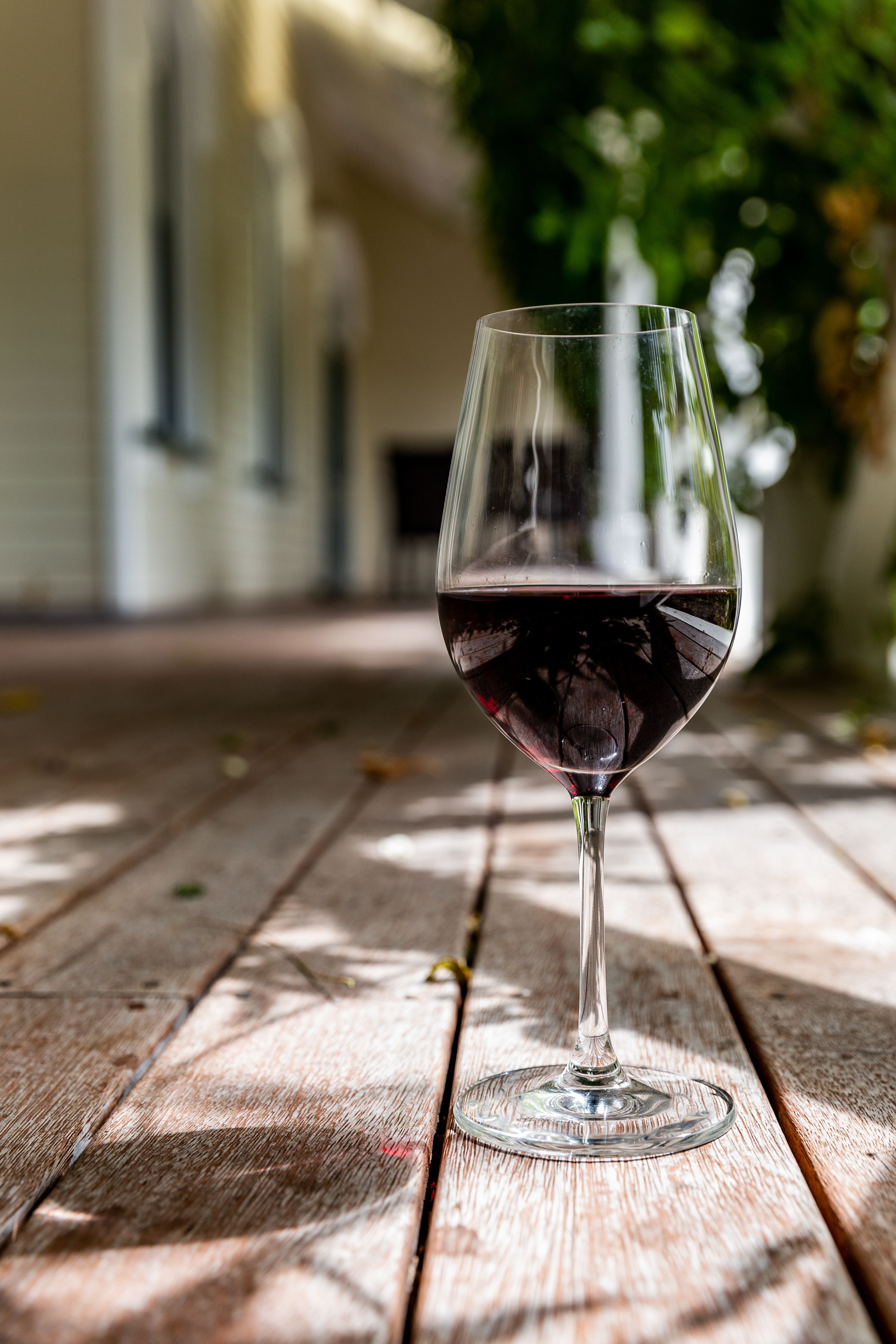 Verre de vin rouge posé sur une terrasse en bois, ombres de feuillage sur les planches