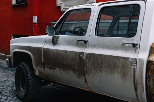 Close-up of a weathered white pickup truck parked near a red wall in an urban area.