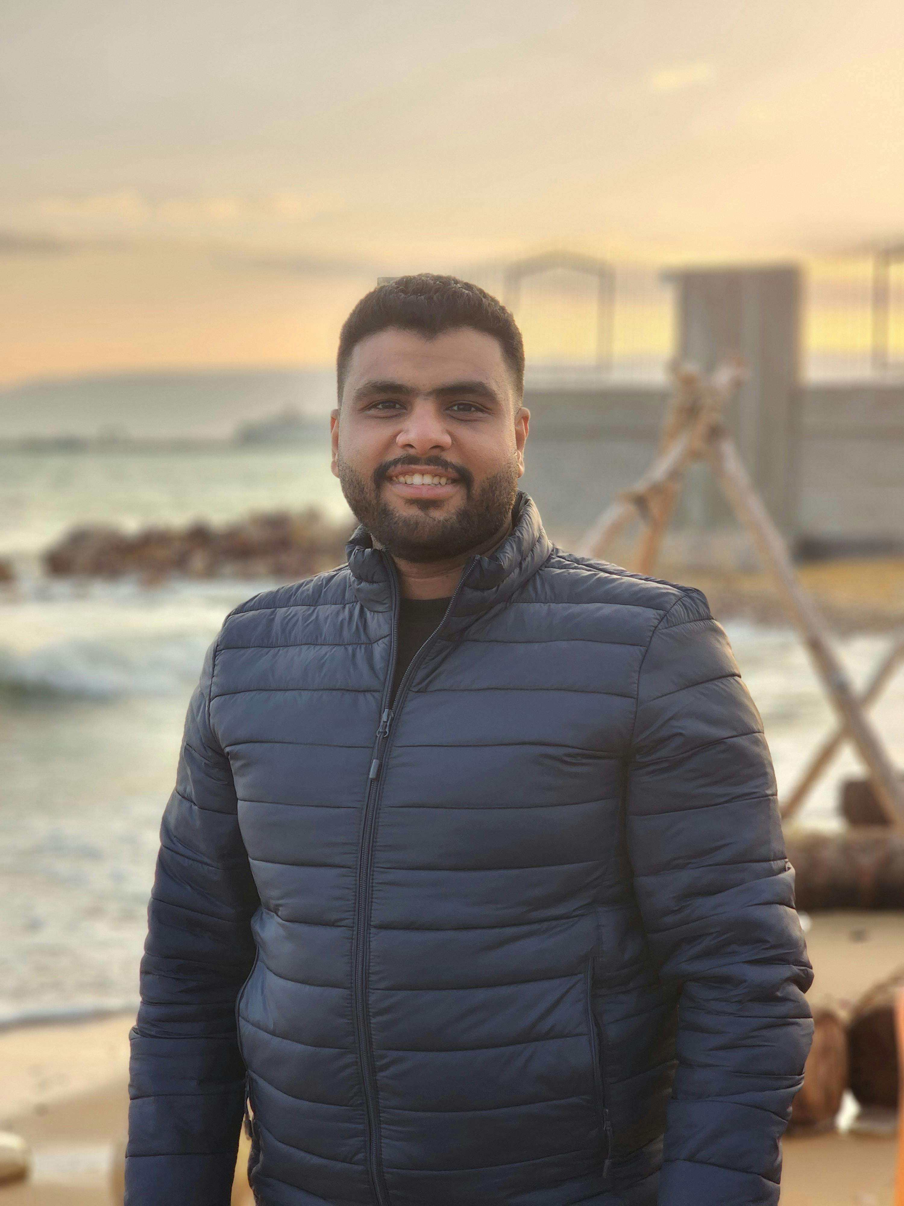 Portrait of a smiling man wearing a black jacket standing on a beach during a calm day.