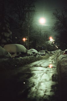 A serene nighttime winter scene with snow-covered cars lining a quiet street under streetlights.