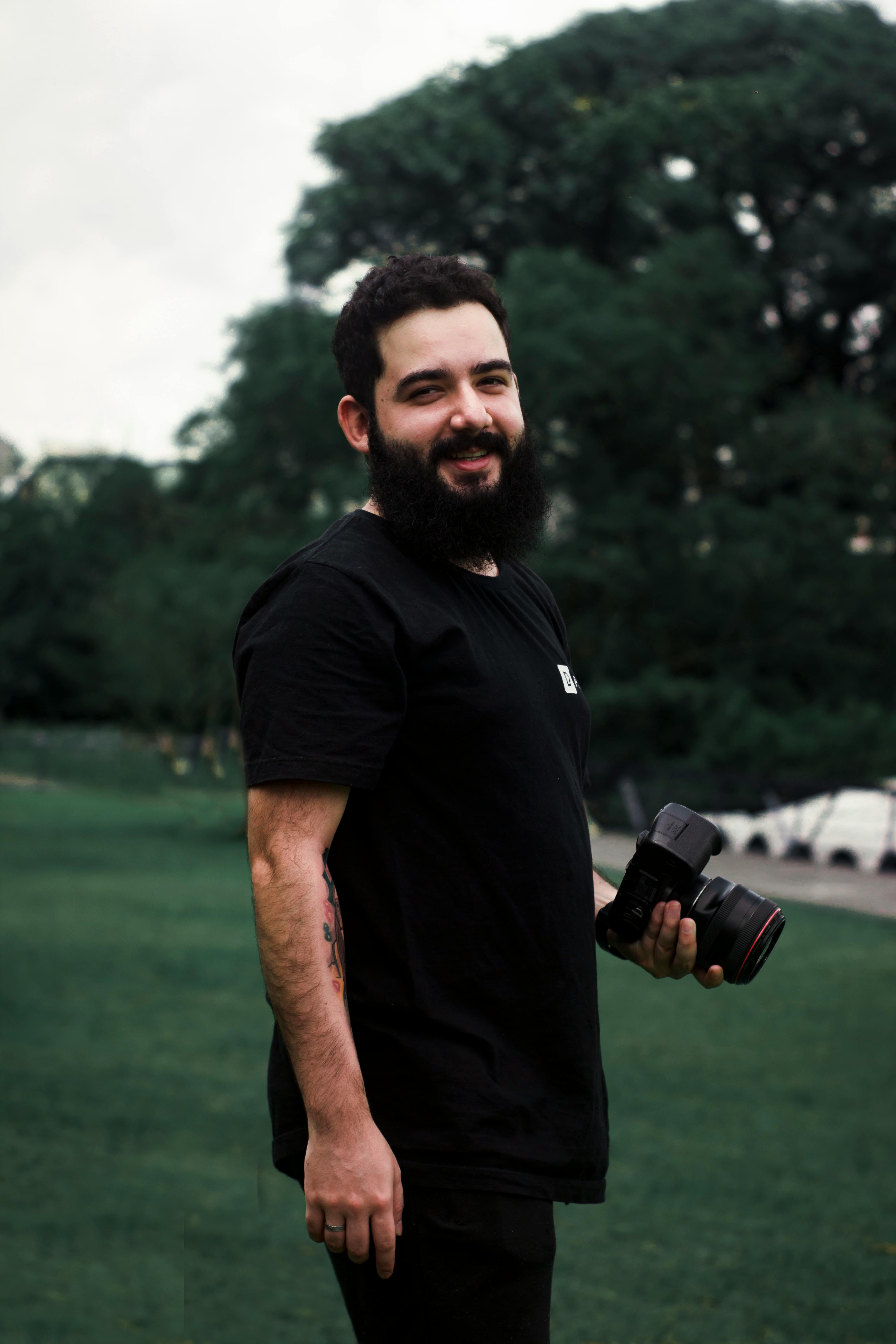 Bearded photographer with camera standing outdoors in a lush park.