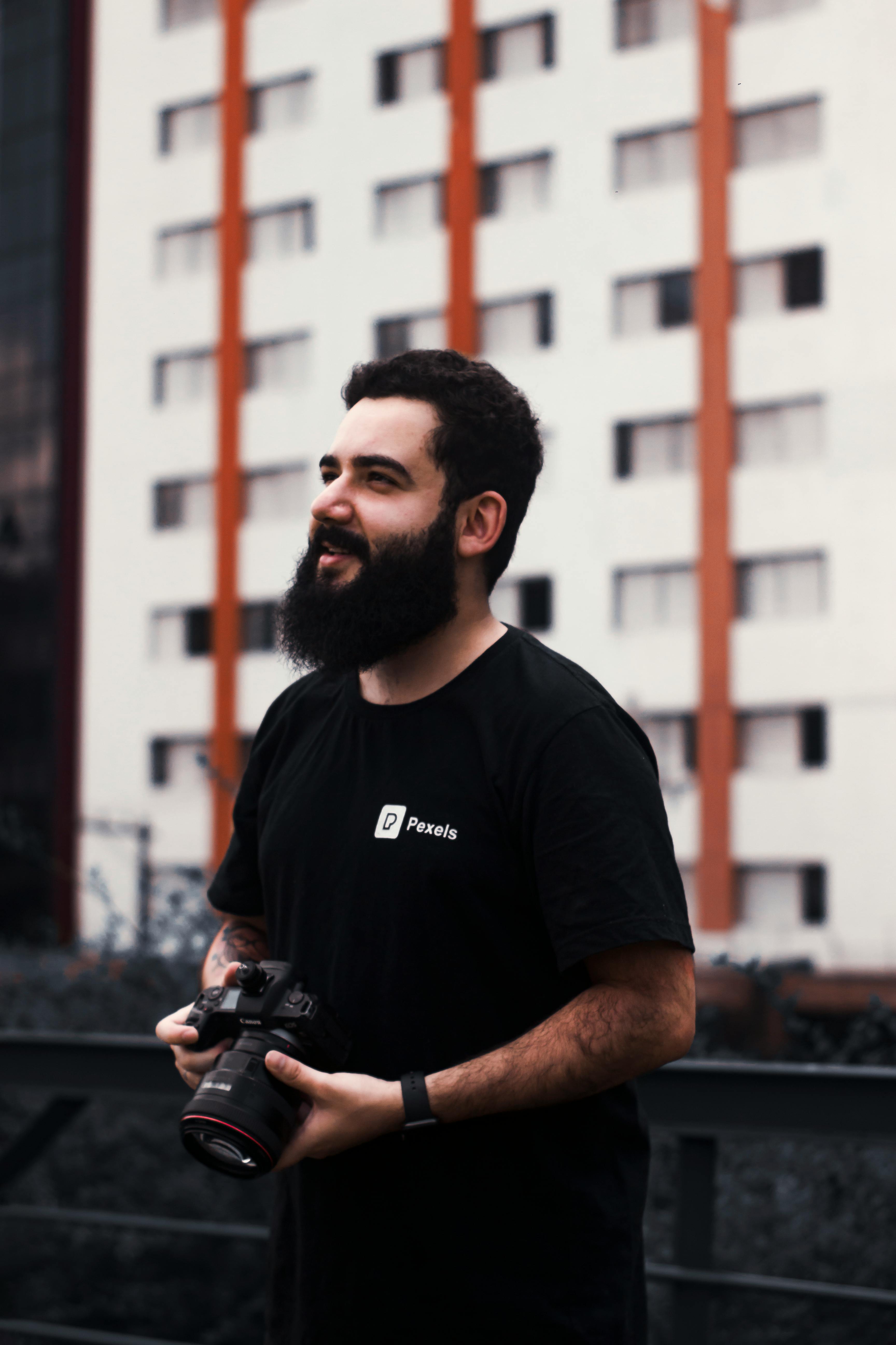 A bearded man holding a camera, standing in front of a city building.