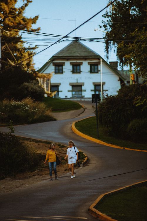Free Two women walking down a curvy road in San Carlos de Bariloche at sunset, surrounded by trees and houses. Stock Photo