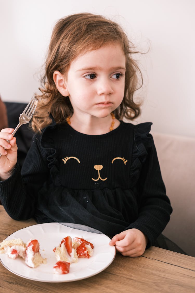 A Cute Little Kid In Black Dress Eating Strawberry Cake While Looking Afar