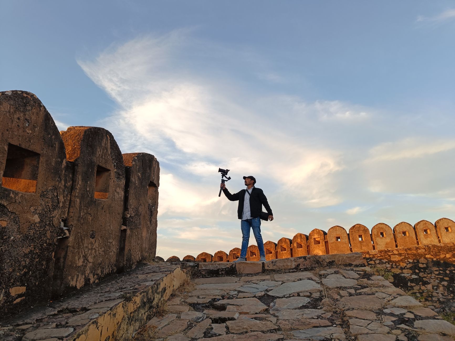 A man capturing video on a fort walkway with a beautiful sky in the background.