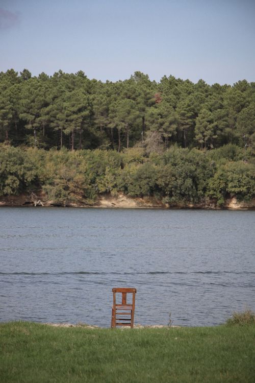 Free A tranquil riverside scene with a single wooden chair, surrounded by lush trees. Stock Photo
