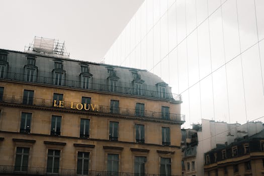 Historic Parisian building reflects on modern glass wall with cloudy sky.