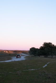 A car travels on a winding road surrounded by fields and trees at dusk.