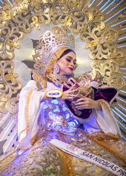 A woman in an ornate costume smiles while holding a decorative doll at a vibrant cultural festival.