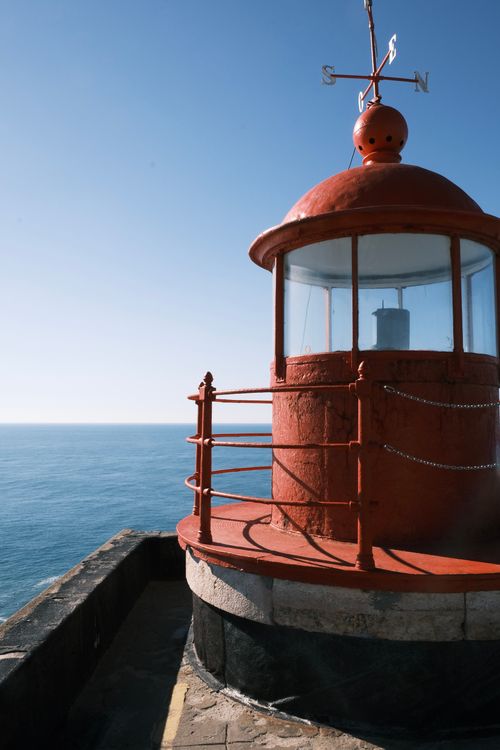 Free A red lighthouse in Nazaré, Portugal stands against a bright blue sky overlooking the Atlantic Ocean. Stock Photo
