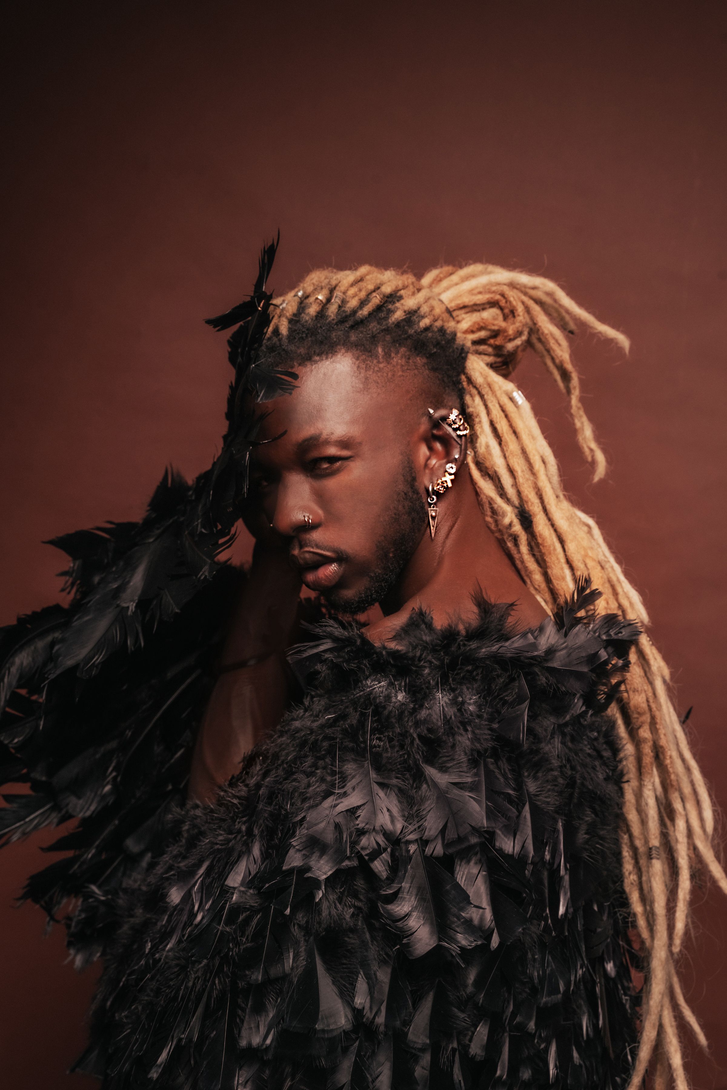 Artistic portrait of a man with long blonde dreadlocks wearing a feathered outfit against a brown backdrop.