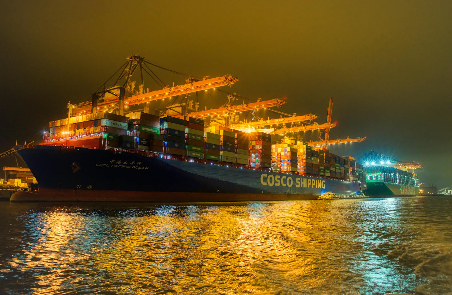 Illuminated container ship and cranes in Rotterdam harbor under overcast night sky.