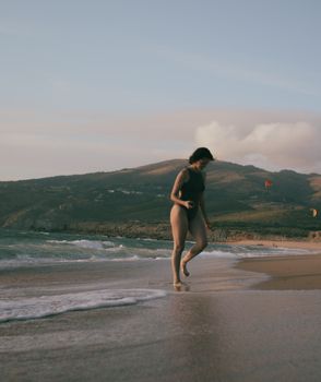 A woman in a black swimsuit walks along a beach with mountains in the background during sunset.
