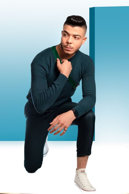 Free Confident young man kneeling in a studio setting, wearing fashionable attire against a blue background. Stock Photo