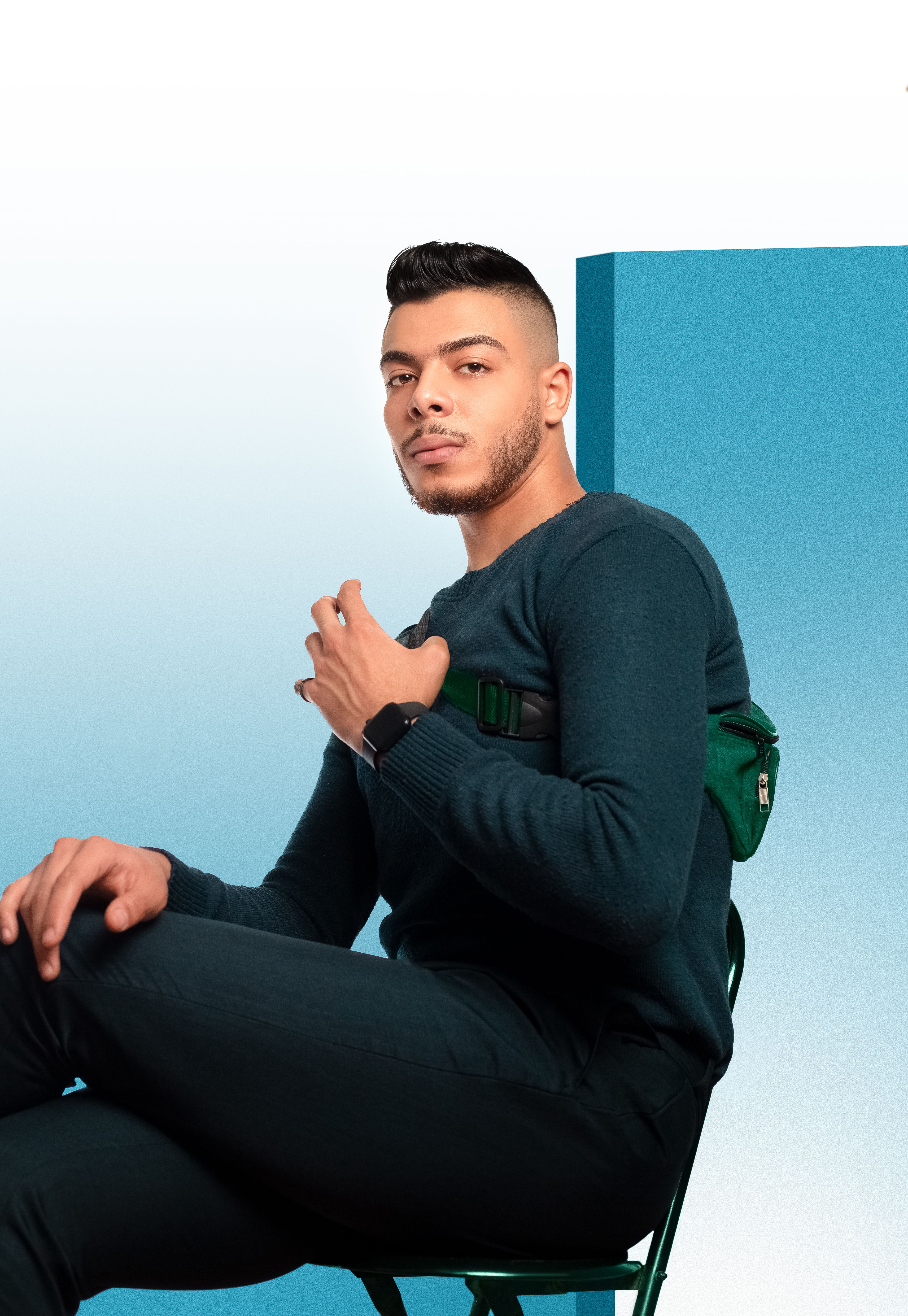 Portrait of a stylish man in a modern outfit sitting confidently against a blue backdrop.