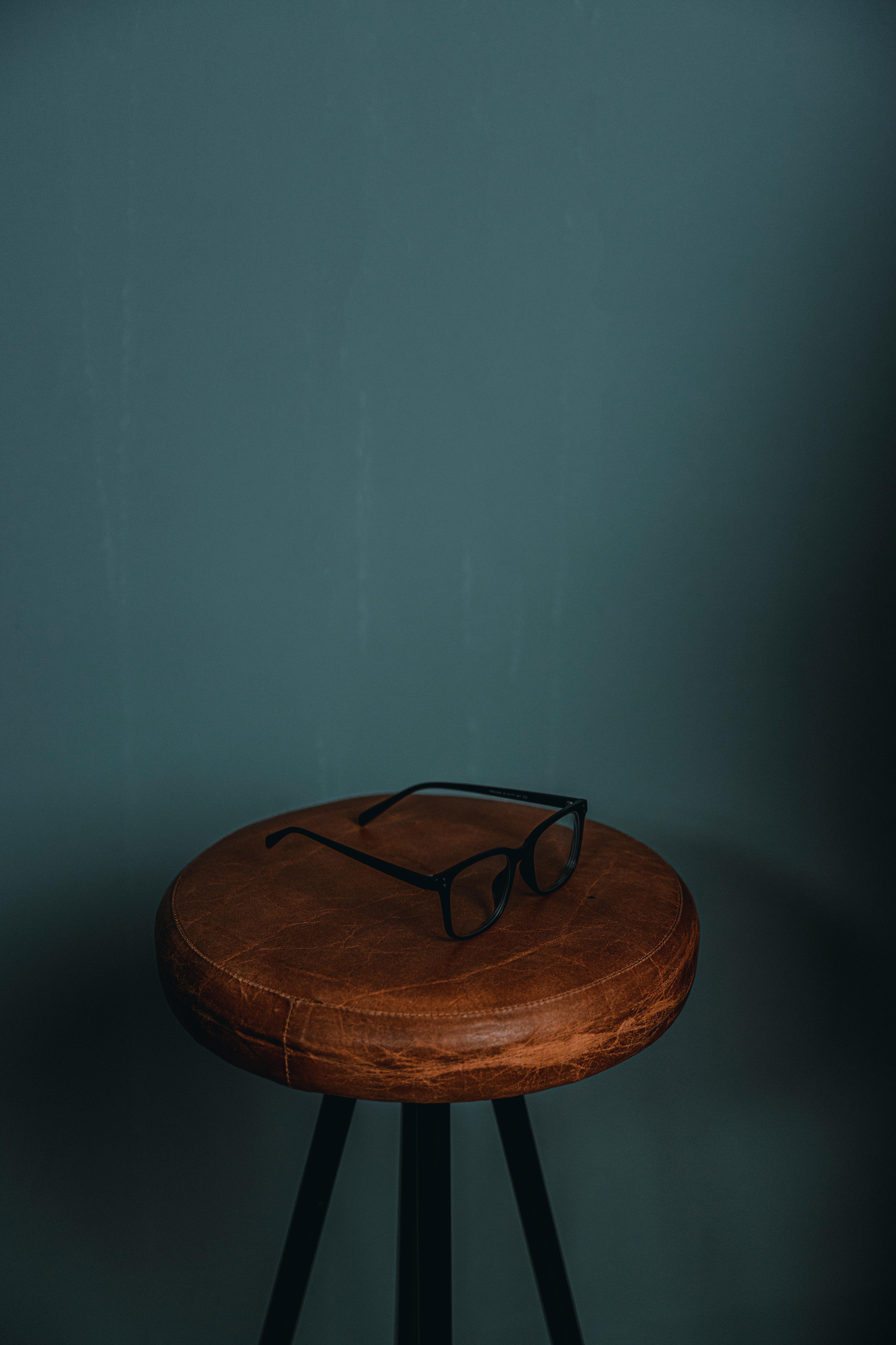 Free Black eyeglasses resting on a brown leather stool against a moody background. Stock Photo