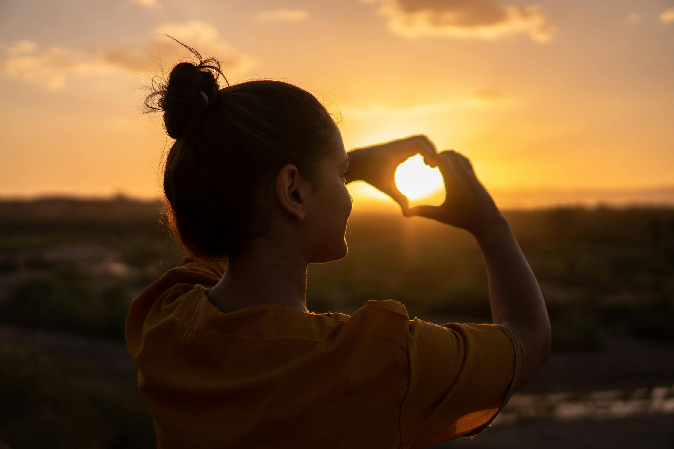 Image of a woman holding her hands in a heart over a sunset. A reminder to love ourselves even if others reject us in dating. 