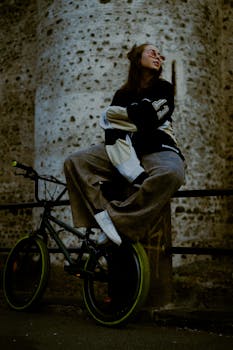Woman in stylish outfit sitting on a bicycle by a stone wall in Catania. Urban lifestyle.