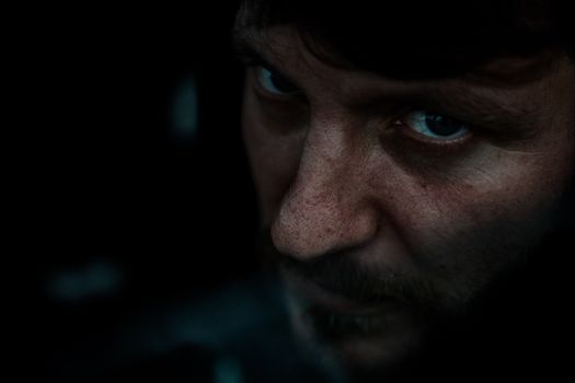 A dramatic close-up portrait of a man with intense eyes and facial features in low light.