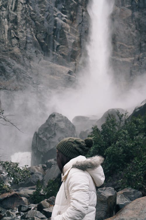Woman in a Jacket and Hat Standing near a Large Waterfall 