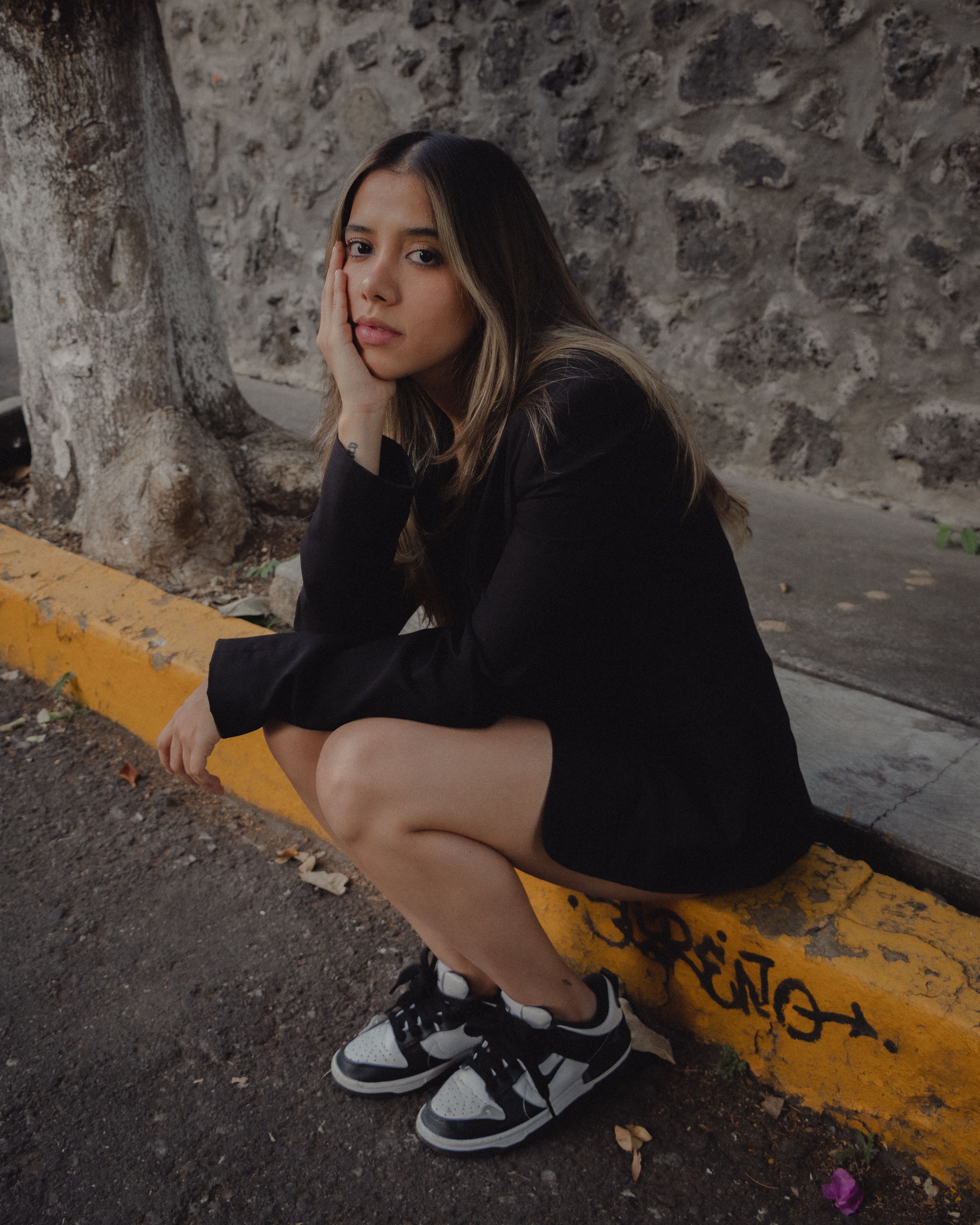 Teen girl in a black outfit sitting thoughtfully on a street curb against a textured stone wall.