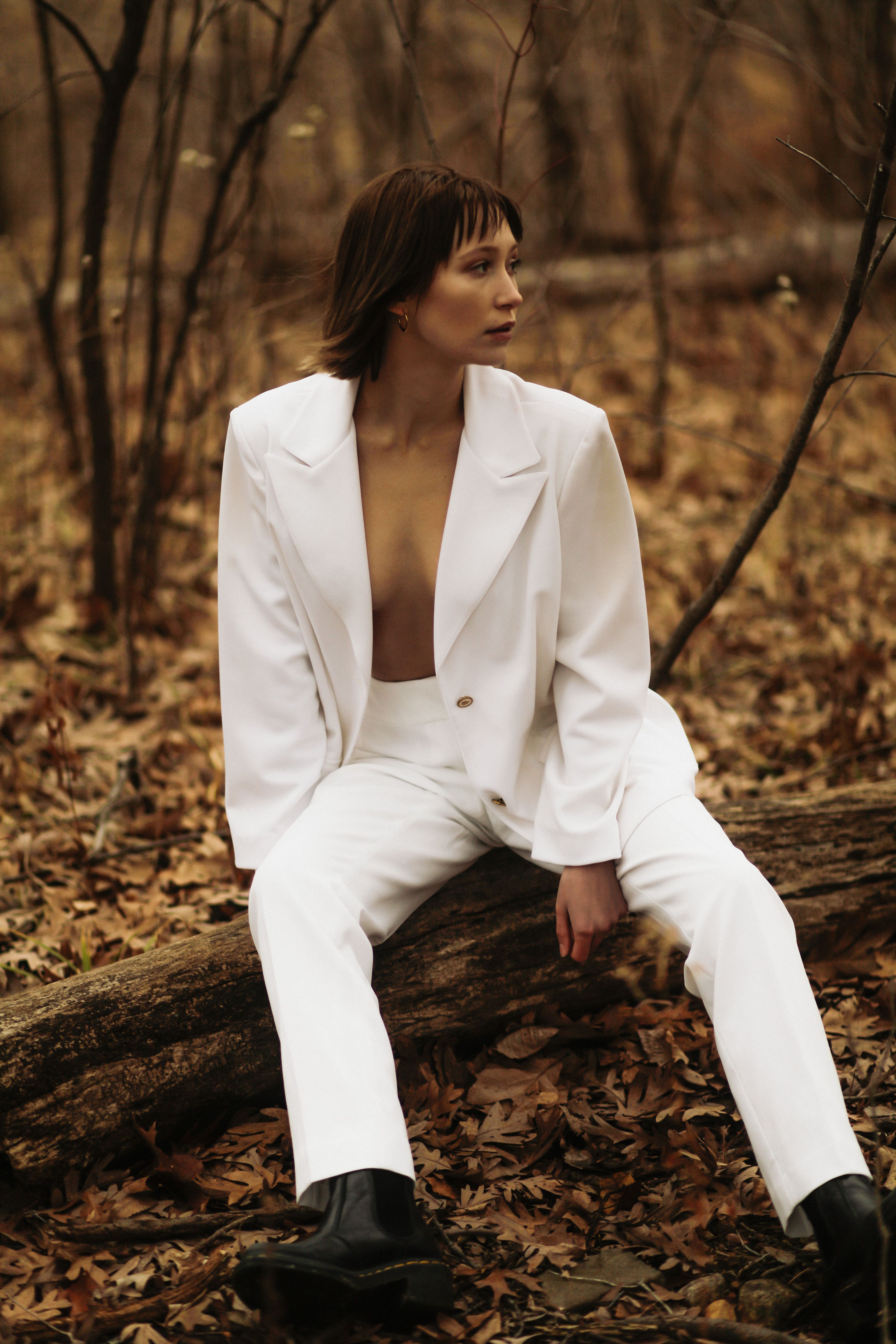 Fashion-forward woman in a white suit seated on a log amid an autumn forest setting.