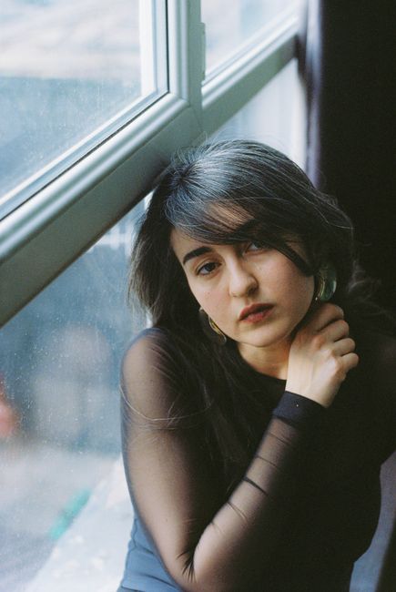 Fashionable woman in a mesh blouse poses thoughtfully by a sunlit window indoors.